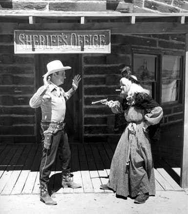 An apparent gag shot as Gene Autry is held up by an old witch on the streets of Pioneertown during the filming of Gene's TV show.