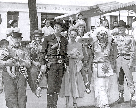In 1948 Hopalong Cassidy took a Western show/rodeo to Honolulu, Hawaii. (Left-Right) Wilbur Plougher with a tiny St. Frances Hospital patient in his arms, Fess Reynolds (Don’s father), Hoppy, unknown, Don Reynolds, stepmother Punkie (above Don), Chief White Eagle, and trick roper Carl Pitti. 