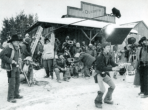 Filming "Silverado" in Santa Fe, NM, gun trainer Arvo Ojala (far left) watches Kevin Costner demonstrate the fast-draw techniques Ojala taught him. Co-star Kevin Kline is at the far right and director Lawrence Kasdan is seated to the left of the camera. 