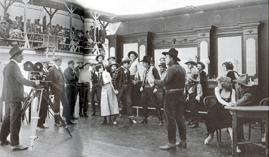 Paying customers in the bleachers watch the filming of “Love’s Lariat” (‘16 Bluebird) starring Harry Carey Sr., seen in the center holding a gun on a badman while protecting Olive Fuller Golden, better known as Carey’s wife. Film was made at Universal’s California studio.