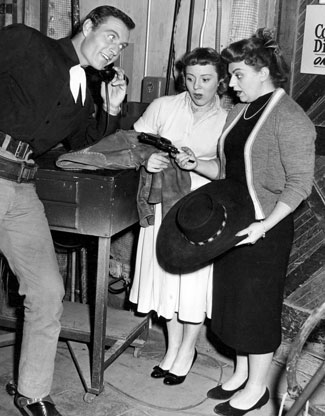 George Montgomery, star of TV’s “Cimarron City” (‘58), talks on the phone while Pat Hitchcock (Alfred’s daughter) and Lorraine Bendix (daughter of William Bendix) look over George’s six-shooter and hat.