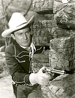 Tex Williams with gun pulled peering from behind rock.